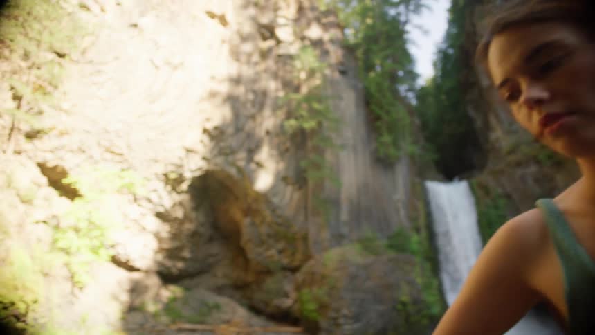 Woman places sunshades on rock with jeans near pond formed by waterfall over cliff