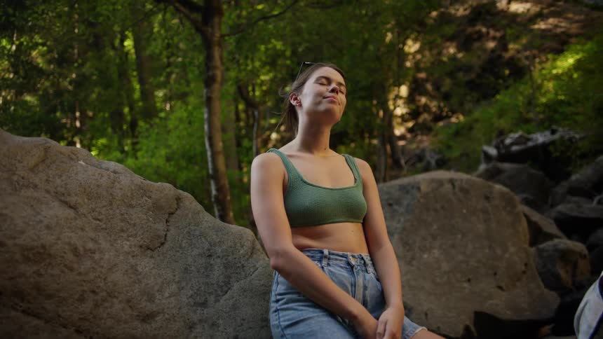 Woman sits against large boulders in forest near incline closes eyes