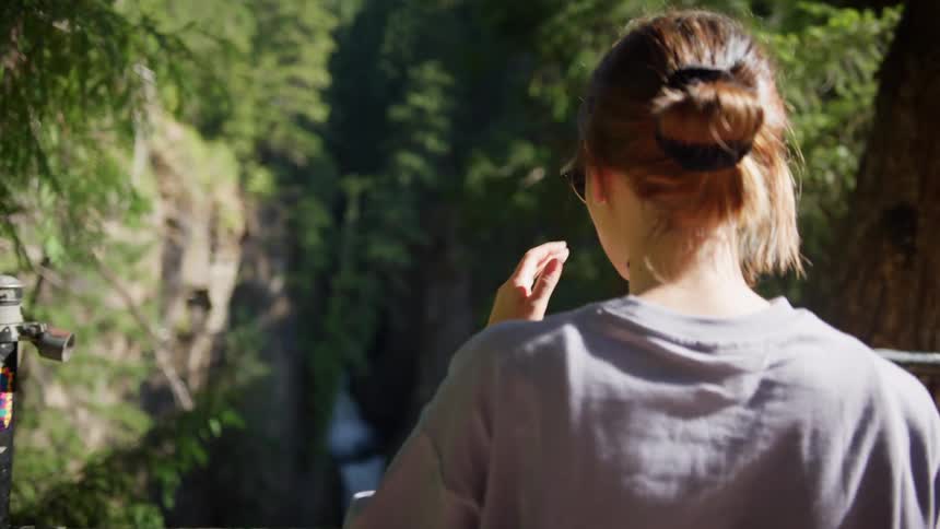 A woman with sunglasses walking next to a fence with trees and a waterfall in the background