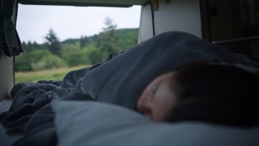 A young woman sleeps in the back of a van parked in a grassland during the day
