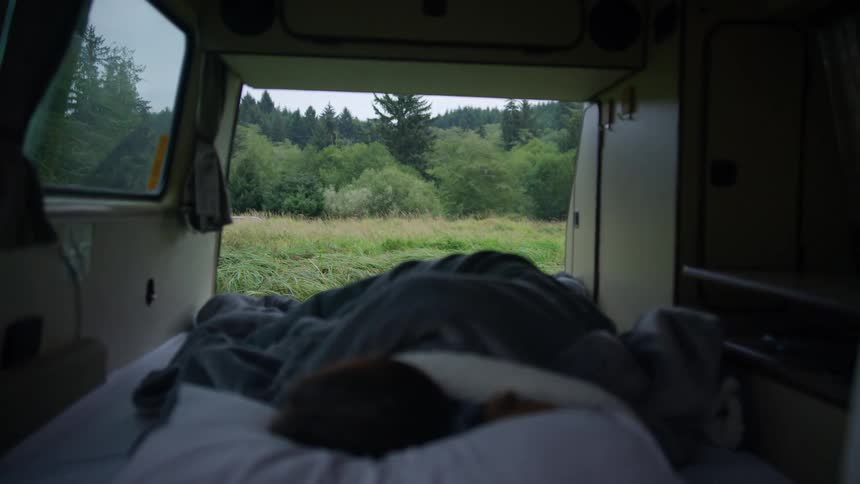 Person sleeps inside caravan with open door showing field and forest
