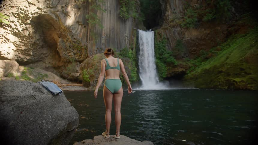 A woman in her underwear steps into natural pool below a waterfall in the wilderness
