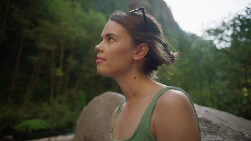 Woman with glasses and green a bikini sits on a rock and watches the waterfall