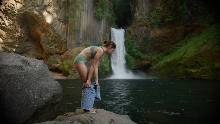 Woman standing on a rock takes off her shorts next to the lake and river