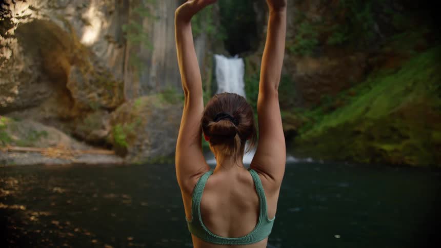 A woman with a bathing suit stretching with a cliff and waterfall in the background