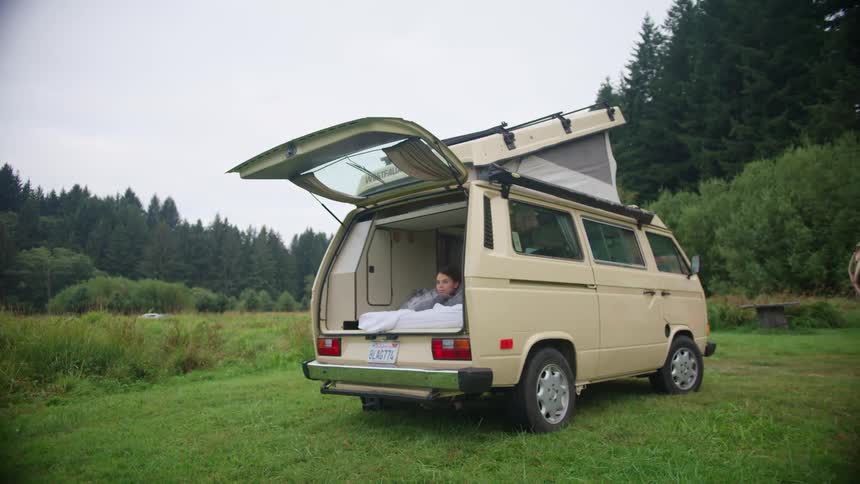 A woman laying down on a bed in a van with trees the sky and clouds in the background