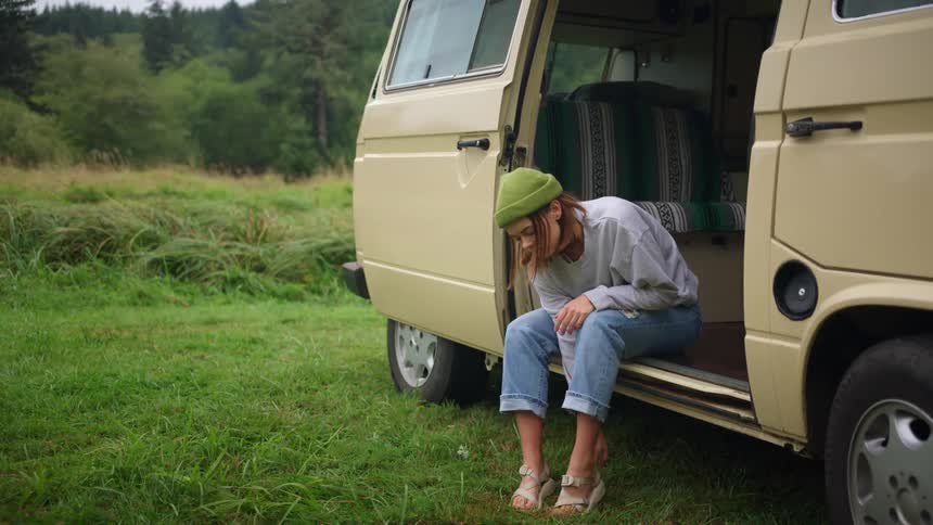 Woman sits on floor board in open van donning shoes near wilderness covered hill