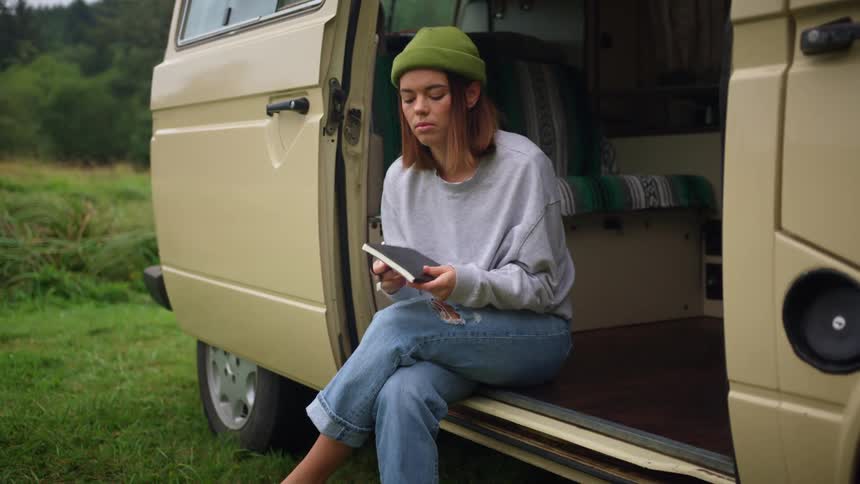 Woman sits on floorboard of van parked in meadow near hill opening journal