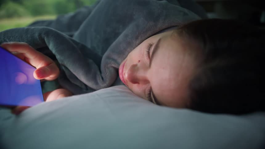 A girl lies on her side in bed covered with a blanket as she scrolls the screen of her cell phone