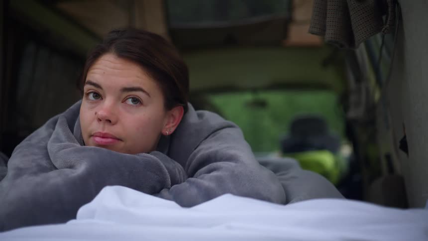 A young woman lies under a blanket in the back of a van during the day