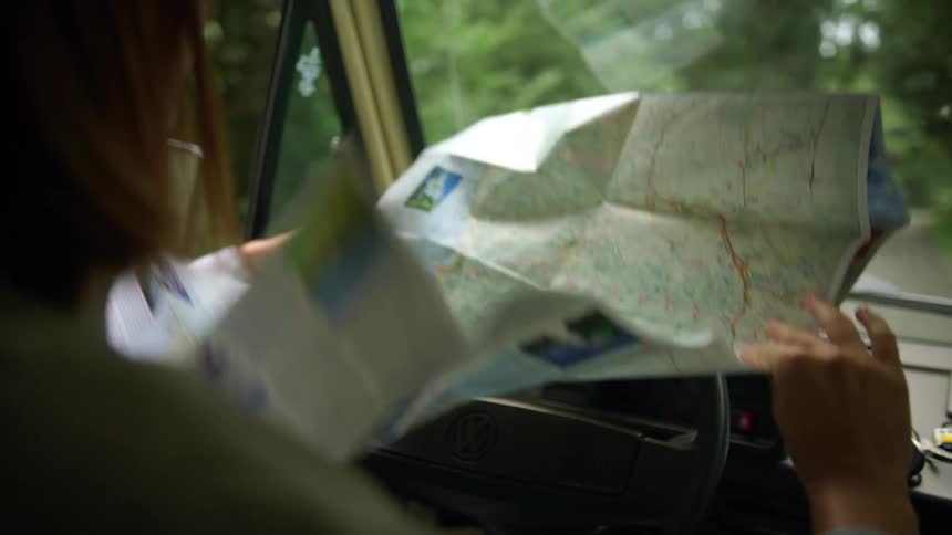 Young woman wearing sweater checks a map over the dashboard of a car parked on a rural road