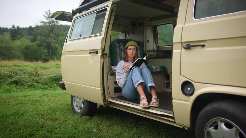 A young woman sits in front of the door in a van parked in a grassland as she writes in a notebook