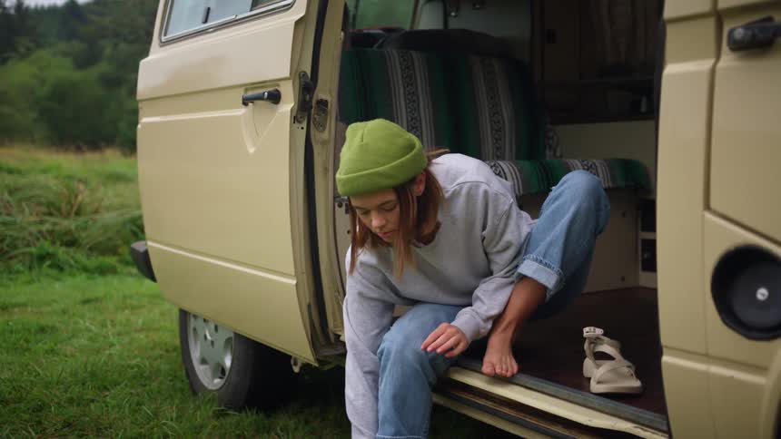 A young woman puts on her sandals while sitting in the doorway of a van parked in a grassland