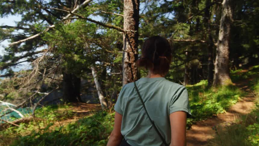 A woman with shades walking on a path in the woods with the sky and clouds in the background