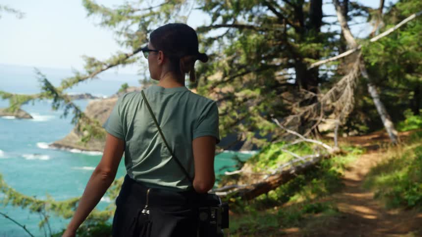 A woman carries a camera as she hikes on a trail on a cliff that overlooks the ocean