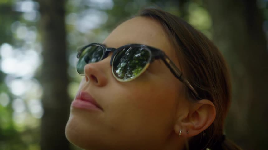 Woman with earrings wears sunglasses and looks up at the trees in the forest