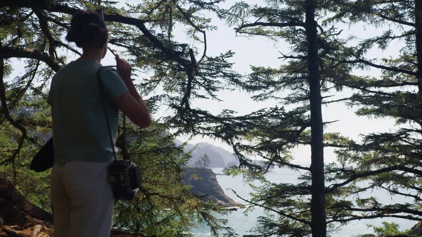 A woman walks to the edge of a cliff in a forest overlooking the ocean and prepares to take a photo with a vintage camera
