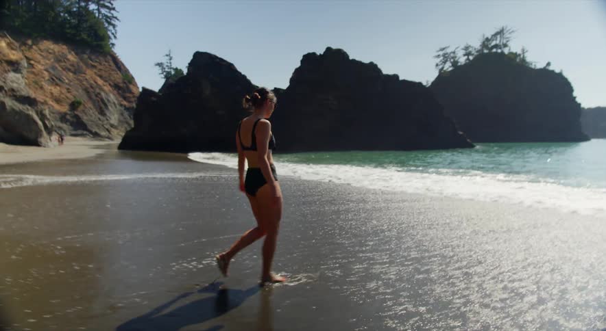 A woman in a black bikini walks on the beach and toward an ocean