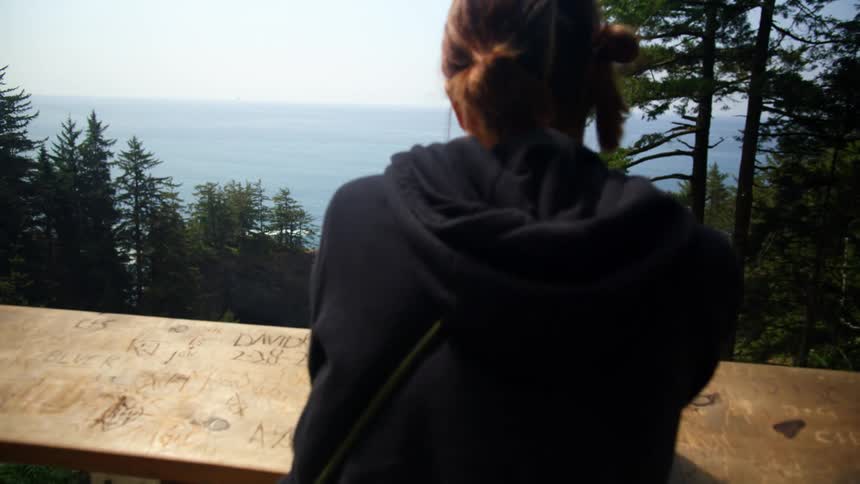 Woman with camera leans against barrier looking toward forest promontory and ocean