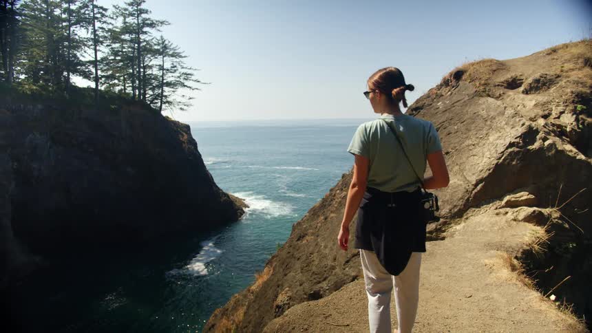 Woman carrying camera walks along narrow path to promontory looking acrosswater to forest covered promontory