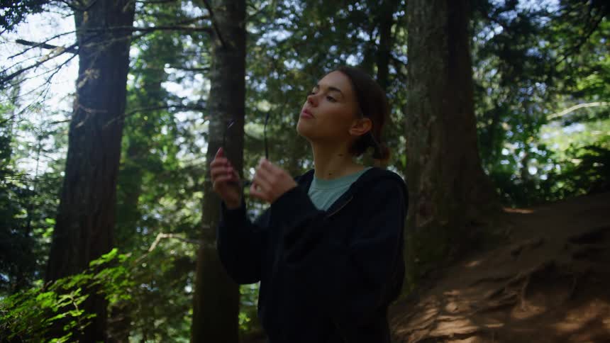 A woman descends a hill in a forest and puts on a pair of sunglasses as she looks up at the sky