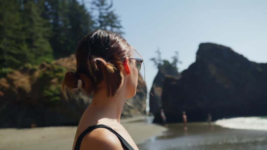 Woman with glasses and a black swimsuit stands and looks at the sea on the beach