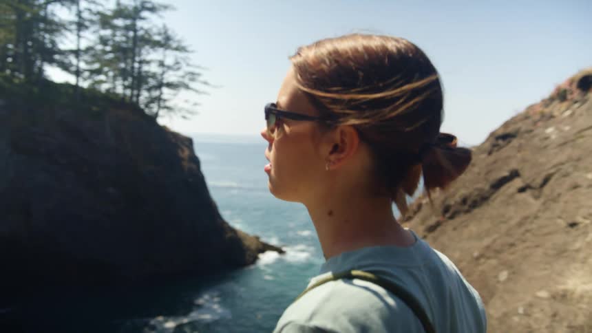 Woman walks along rocky path stopping to view forest covered promontory and ocean