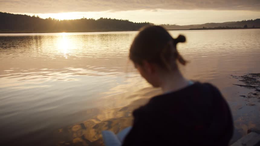 Woman earrings and a zip hoodie reads a book next to the lake at sunset