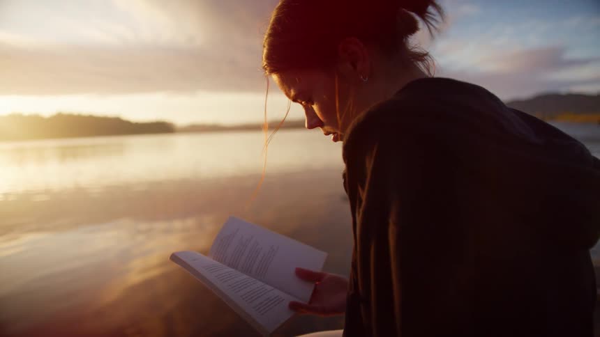 A woman sits by a lake reading a book
