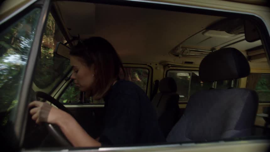 A young woman shifts the gears in a van parked in a woodland during the day