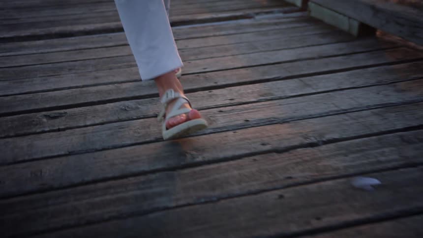 A woman in capris and sandals walks along a boardwalk on a beach at sunset