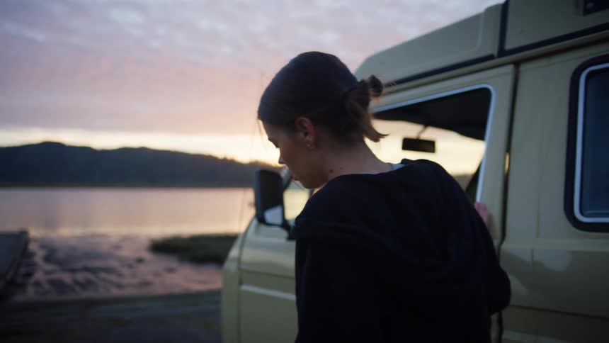 A woman retrieves a book from a van before closing the door and walking towards the shore of a lake in the evening