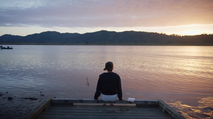 A young woman sits on a wooden pier on a lake a sunset