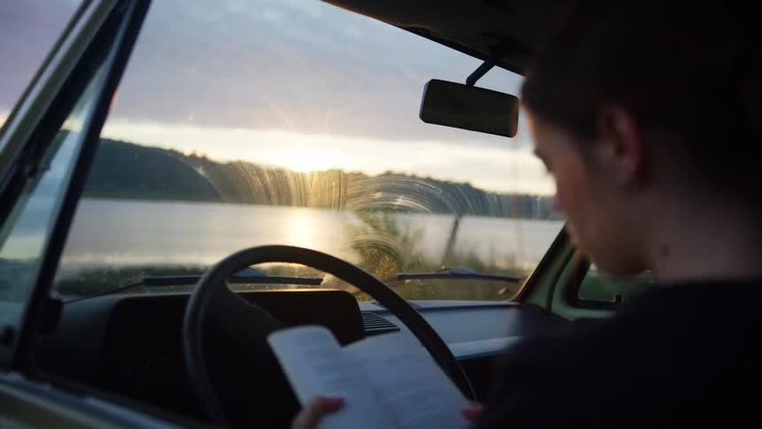 A woman sitting in a vehicle reading a book with the sky and clouds in the background