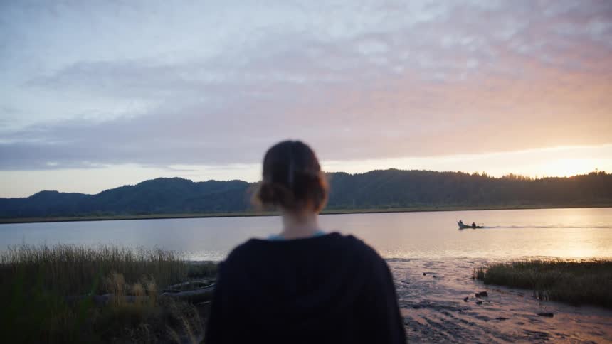A woman walking on a pier towards a river with the sky and clouds in the background