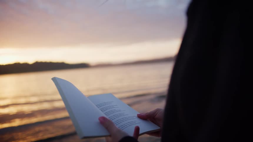 Woman earrings and a zip hoodie reads a book next to the lake at sunset