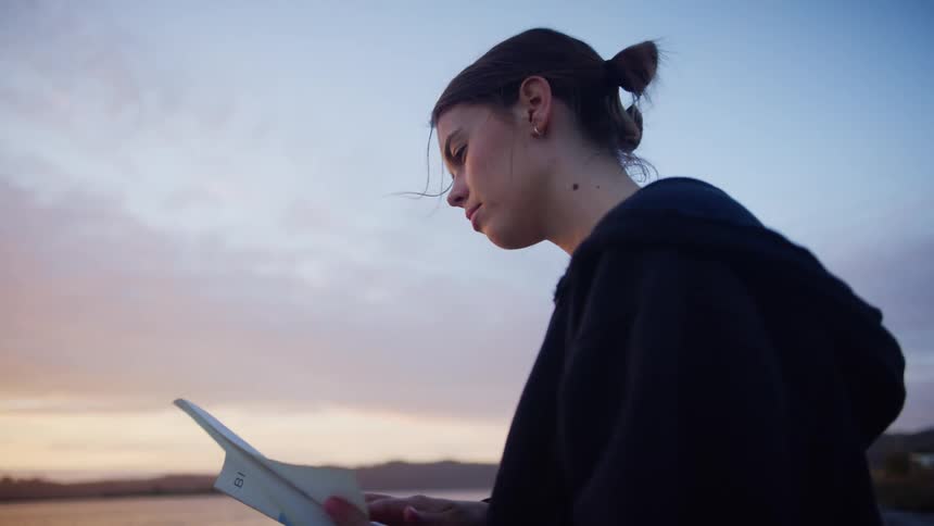 Woman with earrings and a zip hoodie reads a book on the shore at dusk