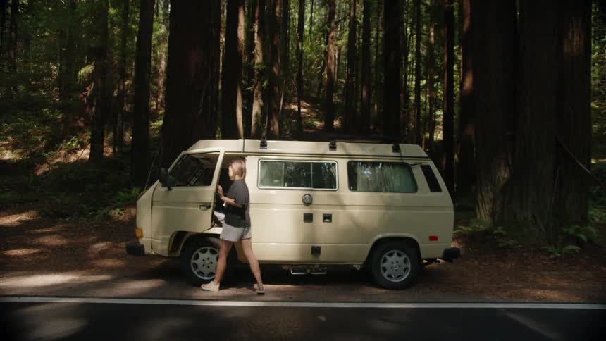 Woman walks to drivers side and gets into van parked on edge of road next to tall forest trees
