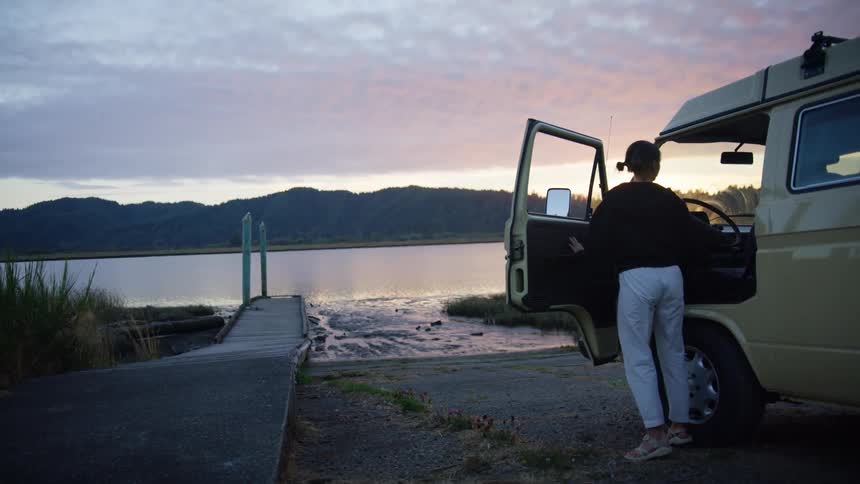 A young woman parked on the beach gets out of her van and grabs a book off the seat