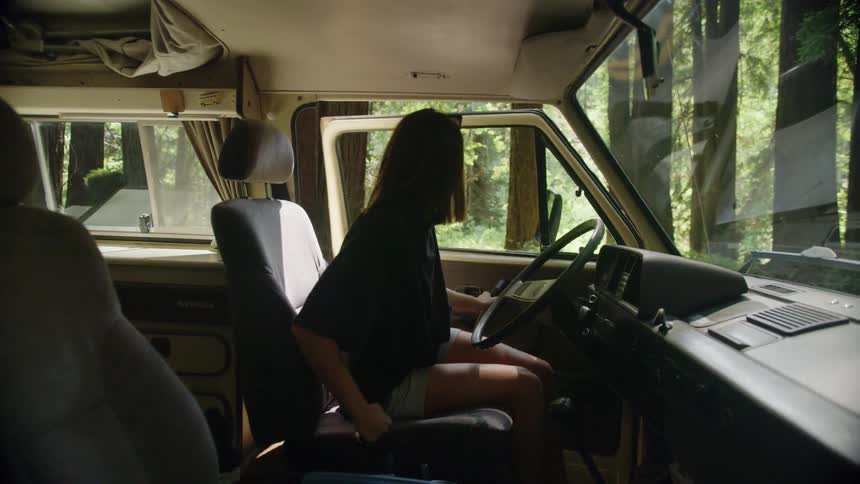 Woman with shorts a black t shirt and brown hair gets in a van and starts the vehicle on the forest road