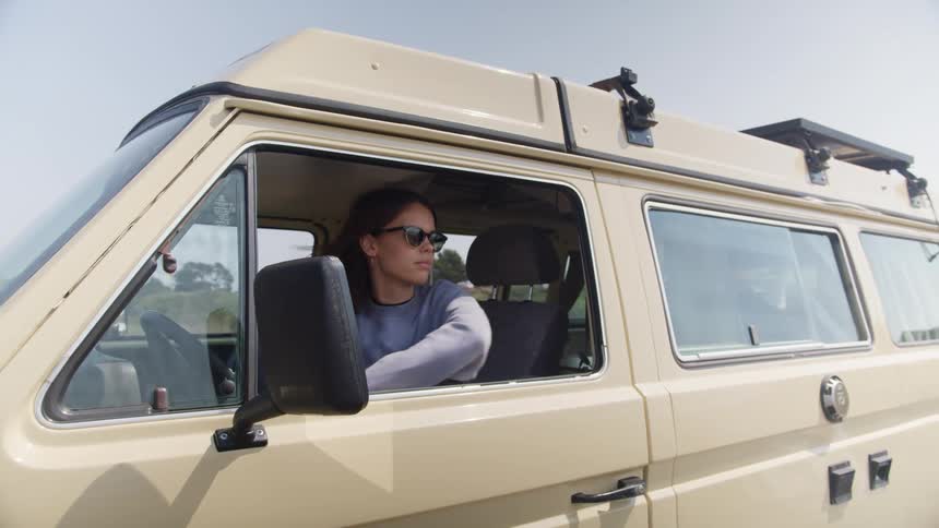 A woman shifts gears in a parked van before turning to rest in the open window while gazing outside