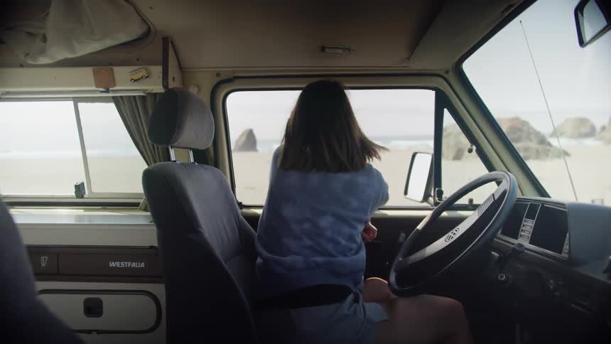 A woman sitting in the drivers seat of a van looks out at the beach on a sunny day