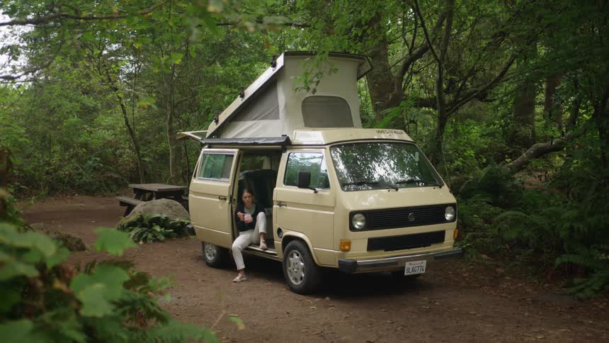 A woman sits and eats in a van turned into a camper thats parked in a forest