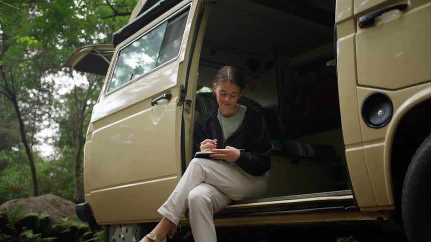 A female sits on the side of an open van and writes in a notebook in a rural setting