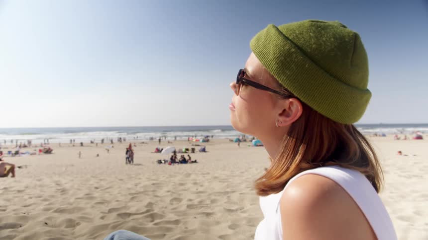 Woman with a green beanie and sunglasses sits on the beach in the sun