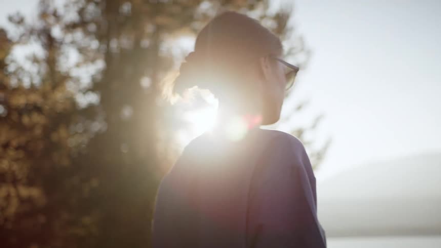 Woman with shorts a blue t shirt and sunglasses wanders around a lake as the sun shines brightly in the background