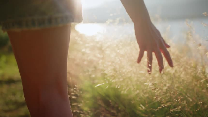 A woman runs her hand through grass as she walks along the shore