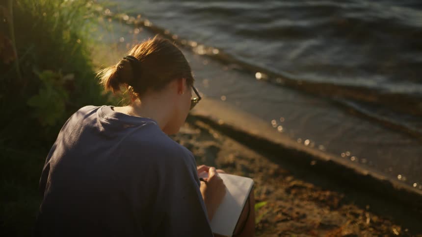 Woman sits on shore writing in notebook as small waves lap nearby