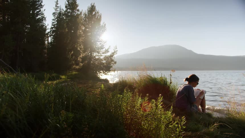 A young woman writes in a notebook while sitting in the grass on the shore of a lake on a sunny day
