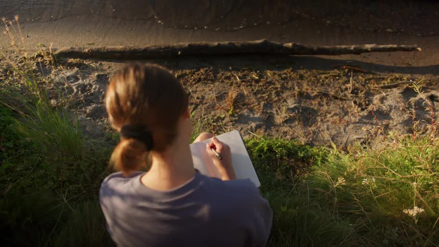 A teenage girl sitting on the shore writes in a notebook